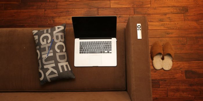 A MacBook on a brown sofa with a cushion beside it.