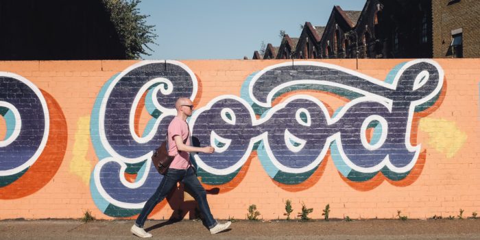 A man walking past a bright orange wall with Good painted on it in huge letters.