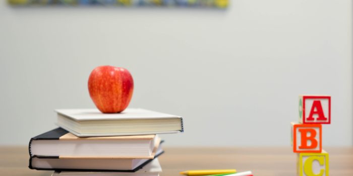 A desk with a pile of books with an apple on top, some pencils and 3 building blocks.